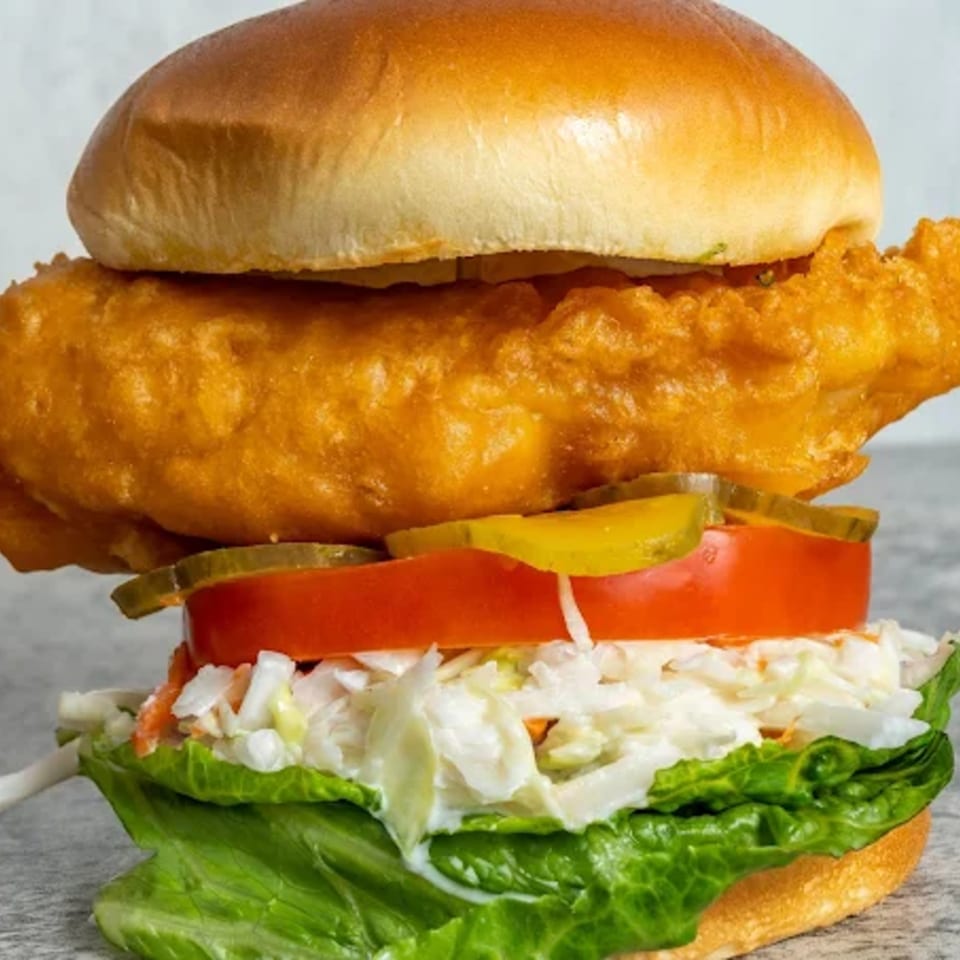 A crispy fried fish sandwich with lettuce, coleslaw, tomato, and pickles on a bun.