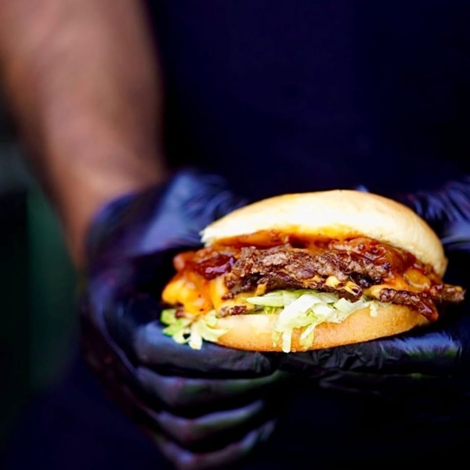 Hands in black gloves holding a Times Square Kitchen American burger with cheese, lettuce, and sauce.