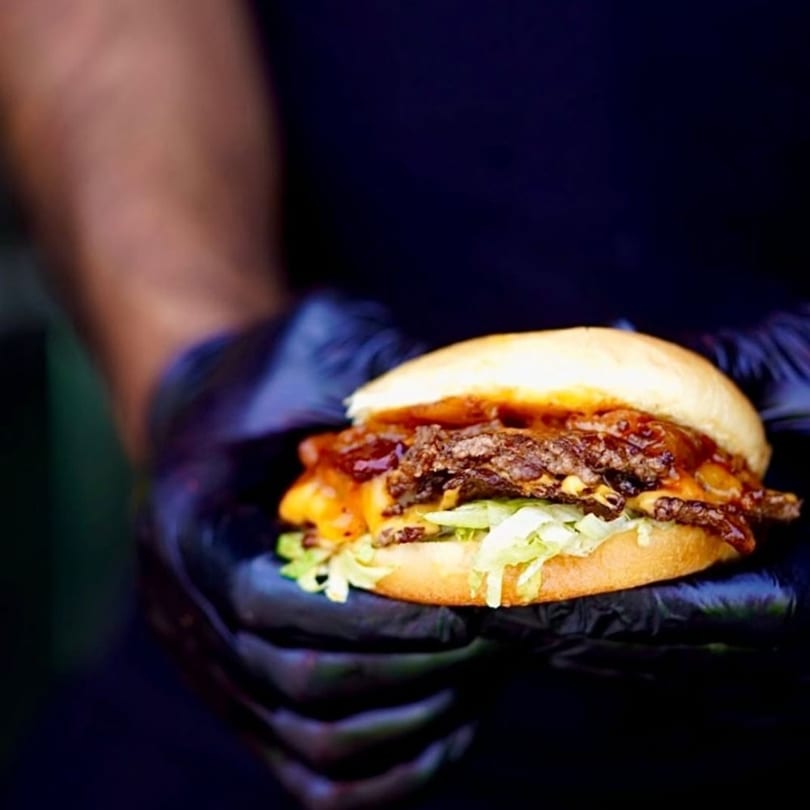 Hands in black gloves holding a Times Square Kitchen American burger with cheese, lettuce, and sauce.